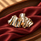 Tie Shop Gold Pleated Ribbon Brooch on red silk. بروش نسائي من تاي شوب مموج باللون الذهبي، إكسسوار أنيق للحجاب والملابس.