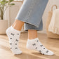 White low-cut socks with a black bow and heart pattern, worn on a model's feet in light-wash jeans, set in a bright, modern room with a wooden floor.