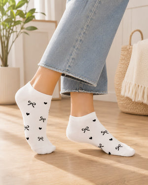 White low-cut socks with a black bow and heart pattern, worn on a model's feet in light-wash jeans, set in a bright, modern room with a wooden floor.
