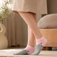 A person wearing a beige dress stands indoors on a rug, showing a pair of low-cut socks. The socks are light pink and heather gray with a small black line-art illustration of a person carrying a baby on their foot.
