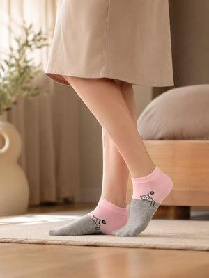 A person wearing a beige dress stands indoors on a rug, showing a pair of low-cut socks. The socks are light pink and heather gray with a small black line-art illustration of a person carrying a baby on their foot.