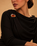 Model wearing a draped black formal dress styled with the Amber Stone Metallic Brooch from Tie Shop on the shoulder for special occasions. موديل ترتدي فستان سهرة أسود منسدل مزين بـ بروش بحجر عنبري إطار معدني من تاي شوب على الكتف للمناسبات.