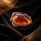 Close-up of Tie Shop Amber Stone Metallic Brooch featuring an amber-colored resin center with grid texture in a textured frame, suitable for abayas and dresses. صورة مقربة لـ بروش بحجر عنبري إطار معدني من تاي شوب بمركز راتنجي عسلي محفور بإطار مموج، مثالي للعبايات والفساتين.