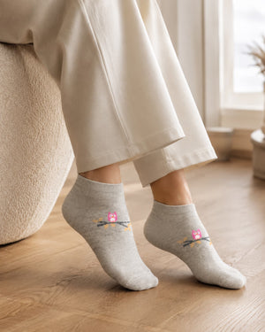 A person wearing light heather gray ankle socks with a small pink owl and orange branch graphic, seated indoors on a wooden floor next to a beige upholstered chair.
