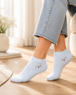 A person wearing light blue ankle socks with a small embroidered cat and red heart design, paired with light blue jeans in a bright indoor setting.