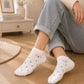 White ankle socks with small black polka dots and a tiny red heart graphic on the toes, worn by a person sitting on a wooden floor in a cozy room setting.