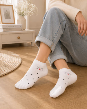 White ankle socks with small black polka dots and a tiny red heart graphic on the toes, worn by a person sitting on a wooden floor in a cozy room setting.