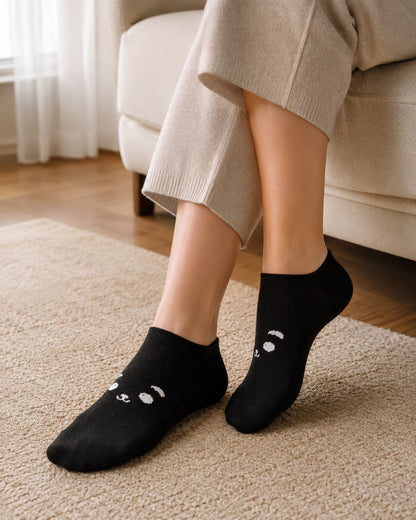 A person wearing black low-cut socks with a white embroidered bear face design, standing on a beige carpet in a living room setting.