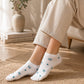 White low-cut socks with small blue bow and heart patterns, worn by a person sitting on a chair in a warm, sunlit room.