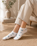 White low-cut socks with small blue bow and heart patterns, worn by a person sitting on a chair in a warm, sunlit room.