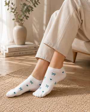 White low-cut socks with small blue bow and heart patterns, worn by a person sitting on a chair in a warm, sunlit room.