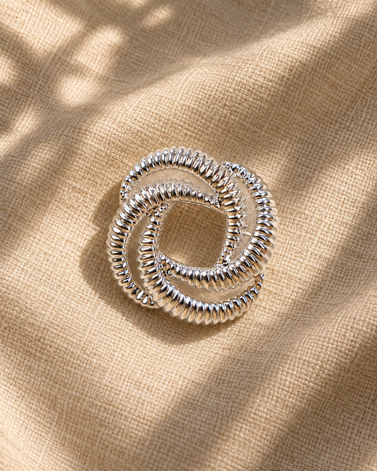 Close-up of Tie Shop Silver Interwoven Knot Brooch on beige fabric. بروش نسائي من تاي شوب بلمسة فضية براقة، إكسسوار أنيق للحجاب والملابس.
