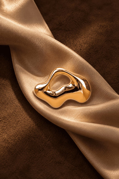 Close-up of Tie Shop Gold Abstract Fluid Brooch on beige silk fabric over brown suede background, a modern shiny metallic pin. صورة مقربة لـ بروش ذهبي لامع بتصميم عضوي انسيابي من تاي شوب على قماش حرير ذهبي وخلفية بني فاخرة، دبوس معدني عصرياكتشفي أناقة بروش تاى شوب مع الحجاب والملابس