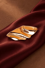 Close-up of Tie Shop Abstract Gold Wave Leaf Brooch displayed on burgundy silk fabric, a modern shiny metallic brooch accessory for women's hijab and clothes. صورة مقربة لـ بروش نسائي من تاي شوب بتصميم ورق شجر ذهبي مموج على قماش حرير عنابي فاخر، إكسسوار أنيق للحجاب والملابس.