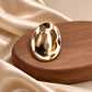 Close-up of Tie Shop Gold Hammered Oval Brooch on wooden base. بروش نسائي من تاي شوب بيضاوي مموج، إكسسوار أنيق للحجاب والملابس.