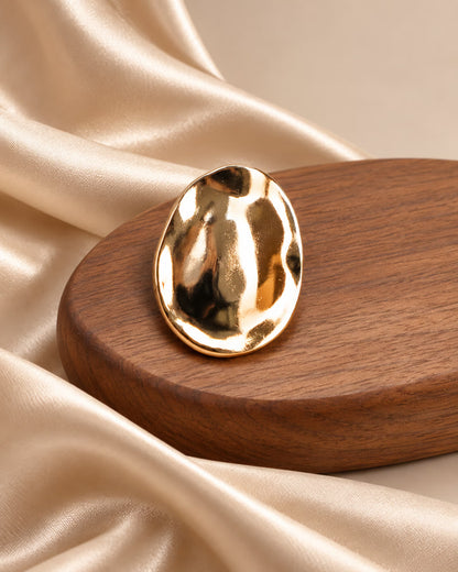 Close-up of Tie Shop Gold Hammered Oval Brooch on wooden base. بروش نسائي من تاي شوب بيضاوي مموج، إكسسوار أنيق للحجاب والملابس.