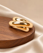 Tie Shop Gold Interlocking Curved Brooch on wooden base. بروش نسائي من تاي شوب بحلقات ذهبية ناعمة ومصقولة، إكسسوار أنيق للحجاب والملابس.