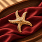 Tie Shop Gold Starfish Brooch on burgundy satin. بروش نسائي من تاي شوب على شكل نجم بحر ذهبي، إكسسوار أنيق للحجاب والملابس.