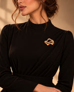Model wearing an elegant draped black dress styled with Tie Shop Gold Crumpled Foil Brooch on the chest, a luxury fashion statement accessory. موديل ترتدي فستان أسود أنيق بياقة ثنيات مزين بـ بروش ذهبي مجعد ومطروق من تاي شوب، لمسة فنية وراقية للملابس اكتشفي أناقة بروش تاى شوب مع الحجاب والملابس
