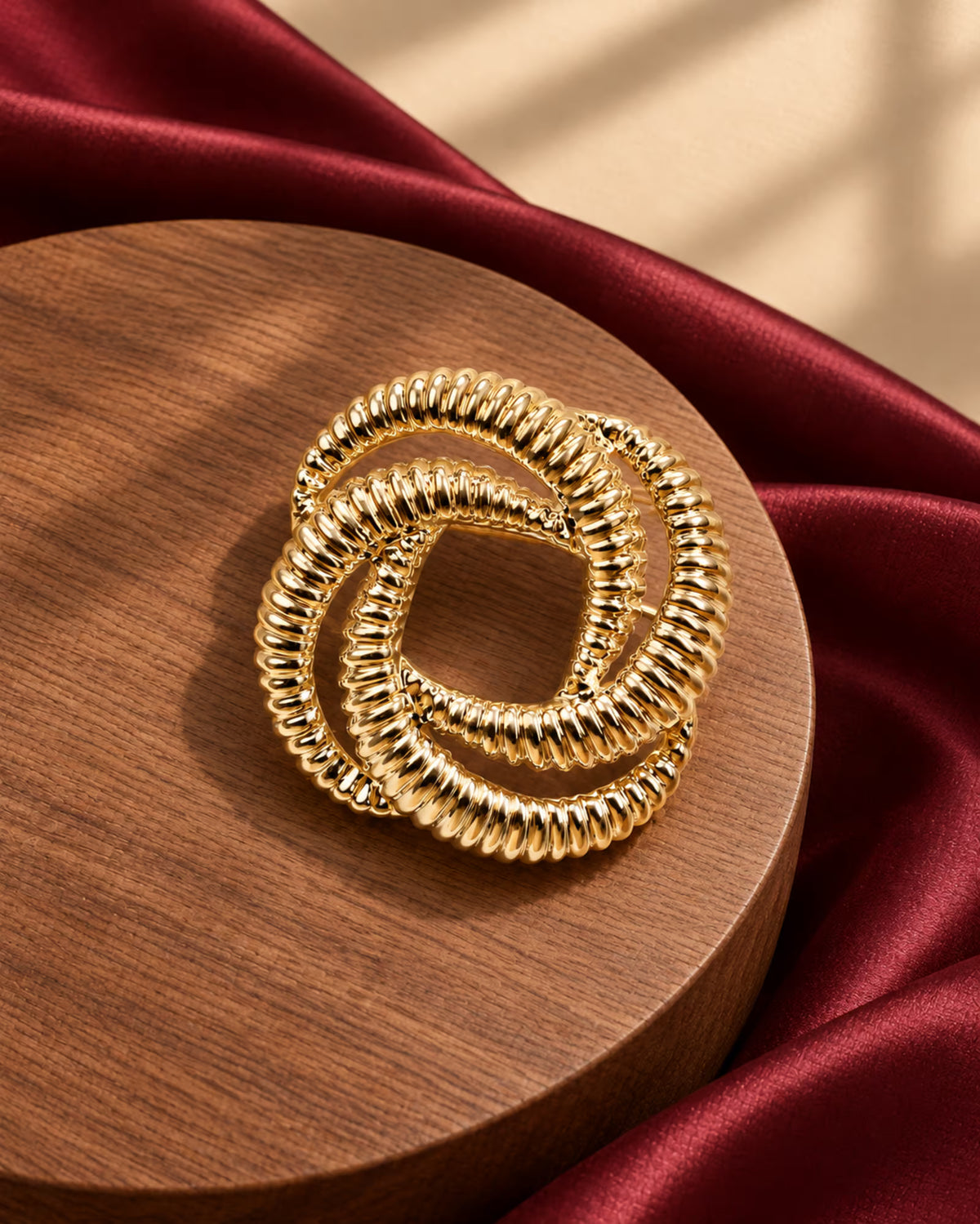 Tie Shop Gold Interlocking Ring Brooch on wooden pedestal with burgundy satin. بروش نسائي من تاي شوب بحلقات ذهبية ملتوية، إكسسوار أنيق للحجاب والملابس.