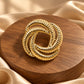Tie Shop Gold Interlocking Ring Brooch on champagne satin background. بروش نسائي من تاي شوب بتصميم عقدة ذهبية مصقولة، إكسسوار أنيق للحجاب والملابس.