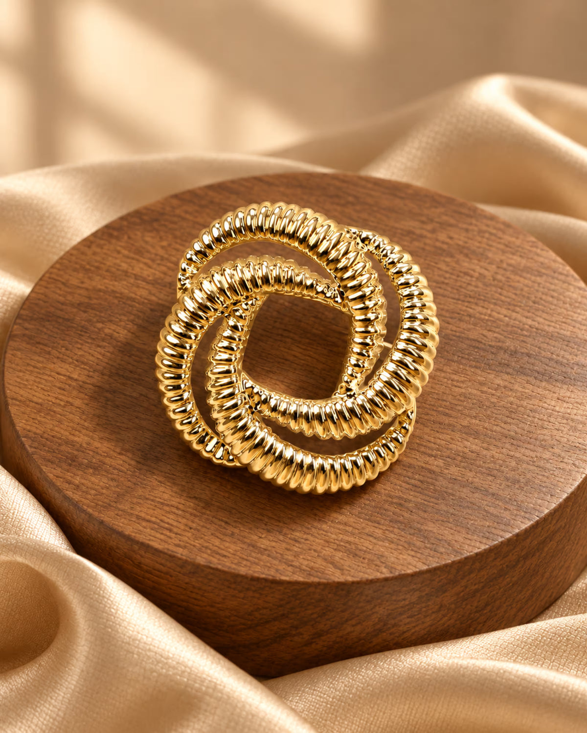 Tie Shop Gold Interlocking Ring Brooch on champagne satin background. بروش نسائي من تاي شوب بتصميم عقدة ذهبية مصقولة، إكسسوار أنيق للحجاب والملابس.