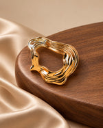 Tie Shop Gold Organic Curved Ring Brooch on wooden base. بروش نسائي من تاي شوب بخطوط مموجة ومصقولة، إكسسوار أنيق للحجاب والملابس.