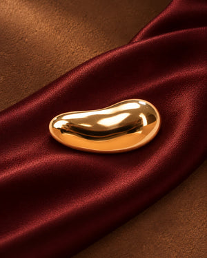 Tie Shop Smooth Gold Bean Brooch on red silk fabric. بروش نسائي من تاي شوب بلمسة ذهبية مصقولة، إكسسوار أنيق للحجاب والملابس.