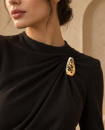 Model wearing an elegant black high-neck gown styled with Tie Shop Gold Textured Crystal Brooch on the chest, a luxury formal brooch. موديل ترتدي فستان أسود أنيق بياقة مرتفعة مزين بـ بروش ذهبي مموج مطعّم بالكريستال من تاي شوب، لمسة فخمة للملابس. اختاري بروش تاى شوب لتنسيق الحجاب والإطلالات