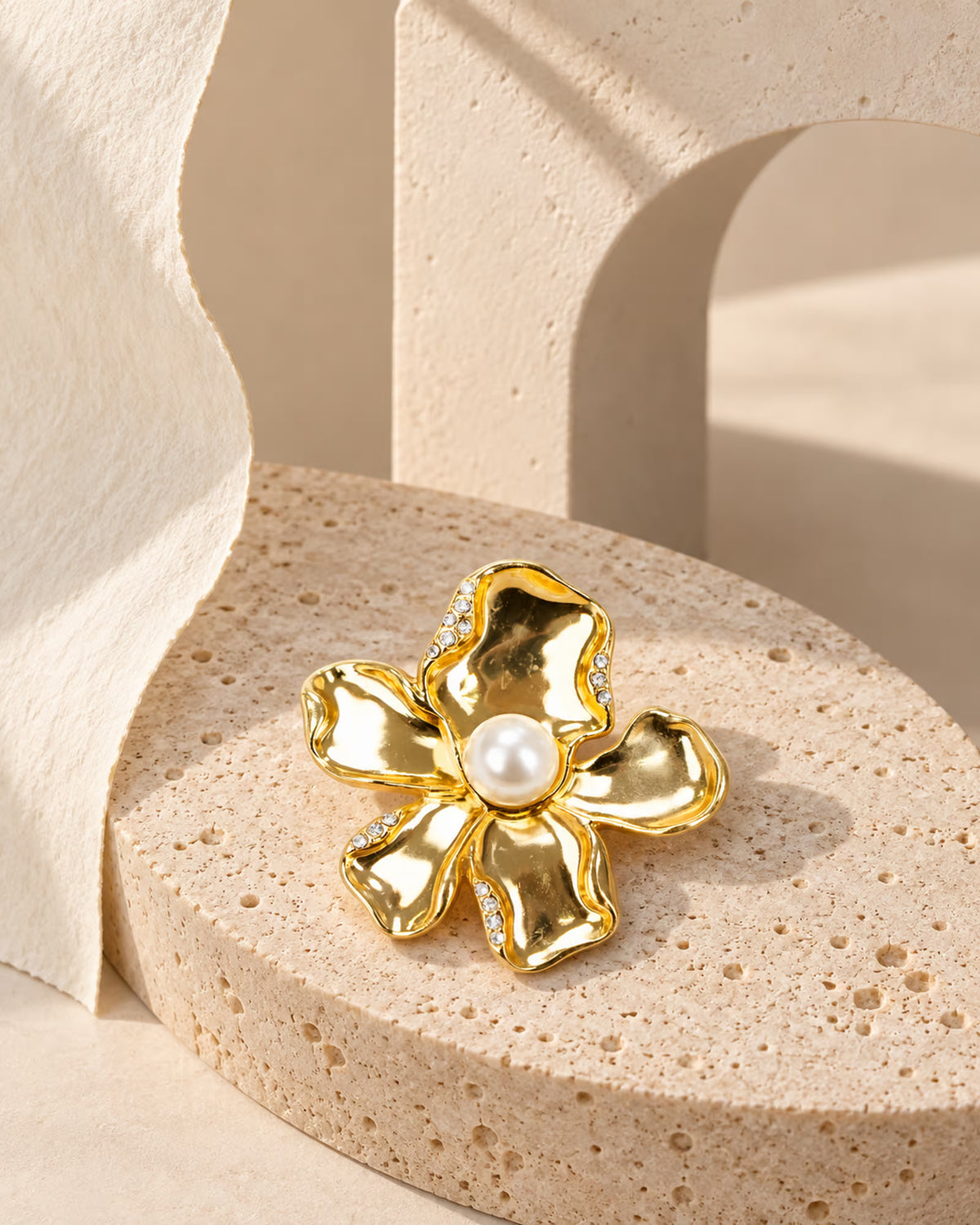 Close-up of Tie Shop Gold Calla Lily Pearl Flower Brooch with crystal accents on a stone pedestal, an elegant pin for abayas and dresses. صورة مقربة لـ بروش وردة اللؤلؤ الذهبي مع تفاصيل الكريستال من تاي شوب على قاعدة حجرية، دبوس أنيق للعبايات والفساتين.