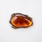 Close-up of Tie Shop Amber Stone Metallic Brooch featuring an amber-colored resin center with grid texture in a textured frame, suitable for abayas and dresses. صورة مقربة لـ بروش بحجر عنبري إطار معدني من تاي شوب بمركز راتنجي عسلي محفور بإطار مموج، مثالي للعبايات والفساتين.