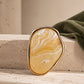 Close-up of Tie Shop Yellow Amber Stone Gold Brooch displayed on a stone surface with olive fabric background, an elegant resin agate brooch accessory for women's hijab and clothes. صورة مقربة لـ بروش نسائي من تاي شوب بتصميم حجر عسلي مموج بإطار ذهبي على قاعدة حجرية، إكسسوار أنيق للحجاب والملابس.