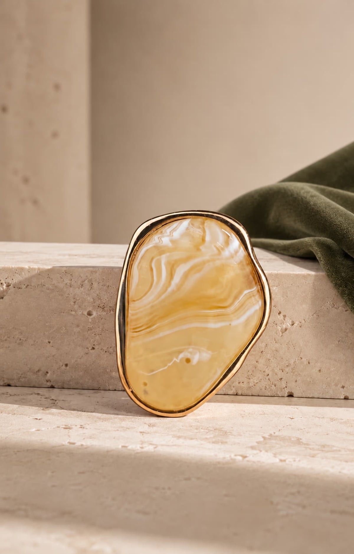 Close-up of Tie Shop Yellow Amber Stone Gold Brooch displayed on a stone surface with olive fabric background, an elegant resin agate brooch accessory for women's hijab and clothes. صورة مقربة لـ بروش نسائي من تاي شوب بتصميم حجر عسلي مموج بإطار ذهبي على قاعدة حجرية، إكسسوار أنيق للحجاب والملابس.