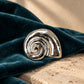 Close-up of Tie Shop Silver Polished Spiral Sea Shell Brooch on dark teal velvet and marble stone, a unique metallic fashion pin. صورة مقربة لـ بروش القوقعة المصقولة الفضي من تاي شوب على قماش مخملي وقاعدة حجرية، دبوس معدني مميز إكسسوارات الحجاب