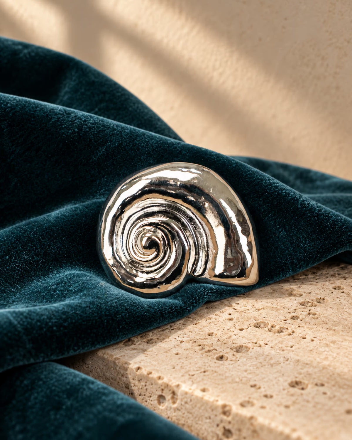 Close-up of Tie Shop Silver Polished Spiral Sea Shell Brooch on dark teal velvet and marble stone, a unique metallic fashion pin. صورة مقربة لـ بروش القوقعة المصقولة الفضي من تاي شوب على قماش مخملي وقاعدة حجرية، دبوس معدني مميز إكسسوارات الحجاب