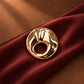 Close-up of Tie Shop Gold Textured Hammered Circle Brooch placed on deep burgundy satin fabric, a luxury gold fashion pin. صورة مقربة لـ بروش دائري ذهبي مموج ومطروق من تاي شوب على قماش ستان عنابي فاخر، دبوس معدني مميز بروش تاى شوب النسائي، إكسسوار أنيق للحجاب والملابس 