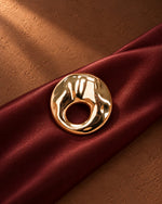 Close-up of Tie Shop Gold Textured Hammered Circle Brooch placed on deep burgundy satin fabric, a luxury gold fashion pin. صورة مقربة لـ بروش دائري ذهبي مموج ومطروق من تاي شوب على قماش ستان عنابي فاخر، دبوس معدني مميز بروش تاى شوب النسائي، إكسسوار أنيق للحجاب والملابس 