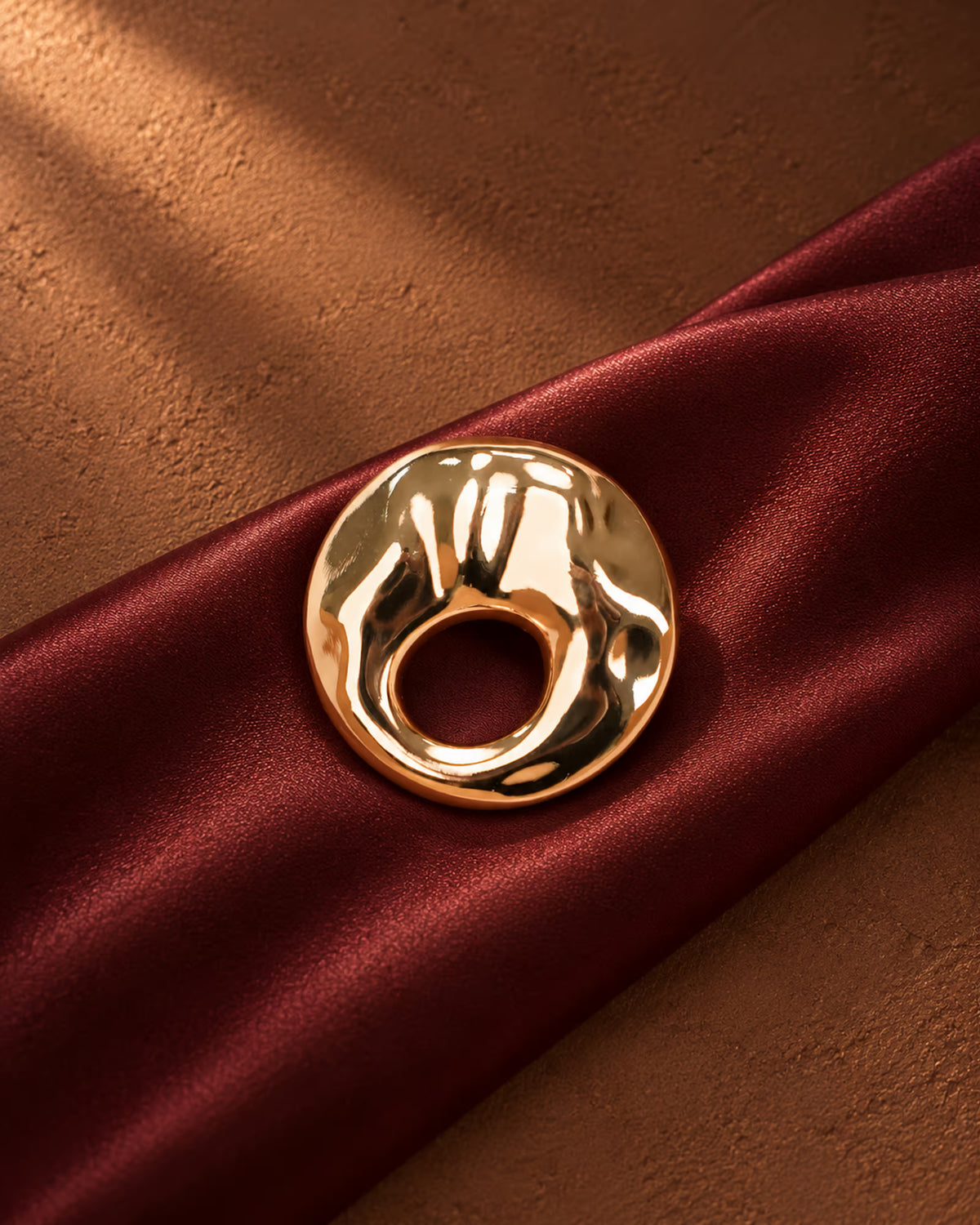 Close-up of Tie Shop Gold Textured Hammered Circle Brooch placed on deep burgundy satin fabric, a luxury gold fashion pin. صورة مقربة لـ بروش دائري ذهبي مموج ومطروق من تاي شوب على قماش ستان عنابي فاخر، دبوس معدني مميز بروش تاى شوب النسائي، إكسسوار أنيق للحجاب والملابس 