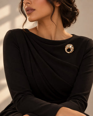 Model wearing a classic black dress styled with Tie Shop Gold Textured Hammered Circle Brooch on the chest, an elegant statement accessory for women. موديل ترتدي فستان أسود أنيق مزين بـ بروش دائري ذهبي مموج ومطروق من تاي  و شوب، اكسسوار راقي وعصري للملابس وللحجاب