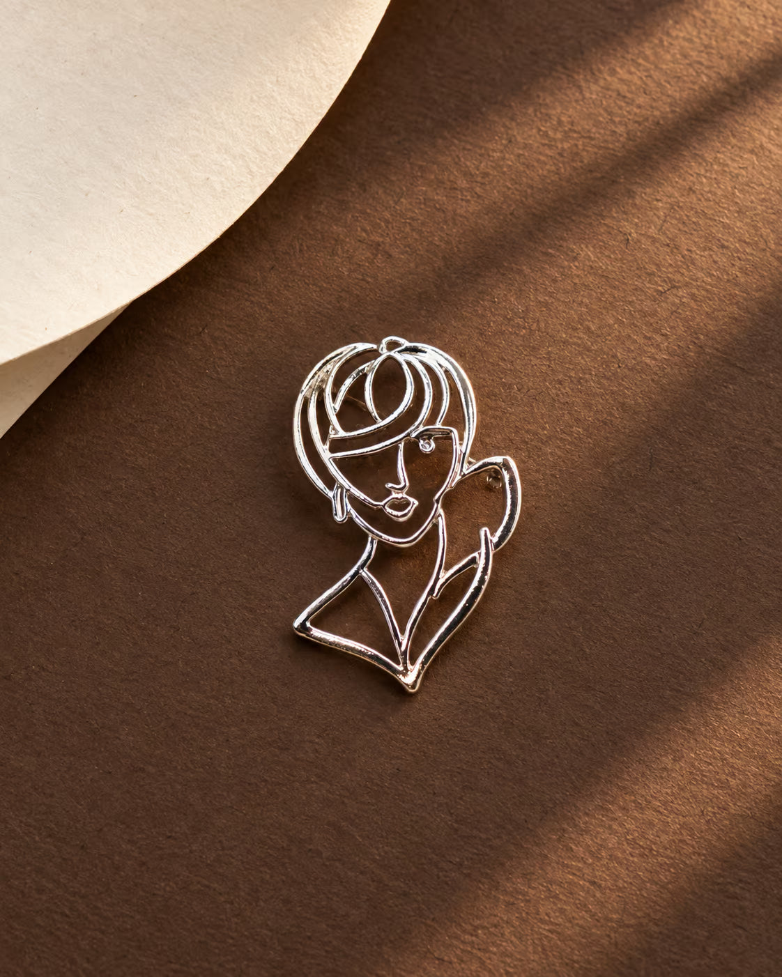 Detail shot of Tie Shop Silver Abstract Line Art Face Brooch on a warm brown surface, an elegant artistic metallic brooch. صورة دقيقة لـ بروش وجه المرأة الفني الفضي من تاي شوب على خلفية بنية دافئة، بروش معدني راقي. إكسسوارات الحجاب من تاى شوب 