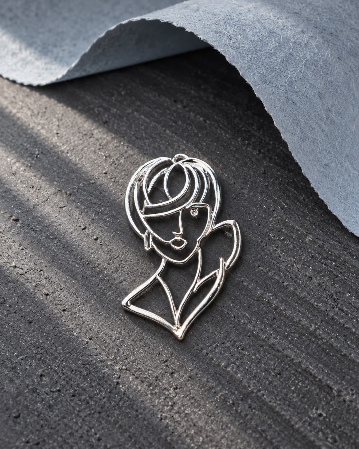 Close-up of Tie Shop Silver Abstract Line Art Face Brooch on a dark textured background with grey felt, a unique aesthetic pin. صورة مقربة لـ بروش وجه المرأة الفني الفضي من تاي شوب على خلفية رمادية، دبوس معدني بتصميم عصري. البروش النسائي من تاى شوب