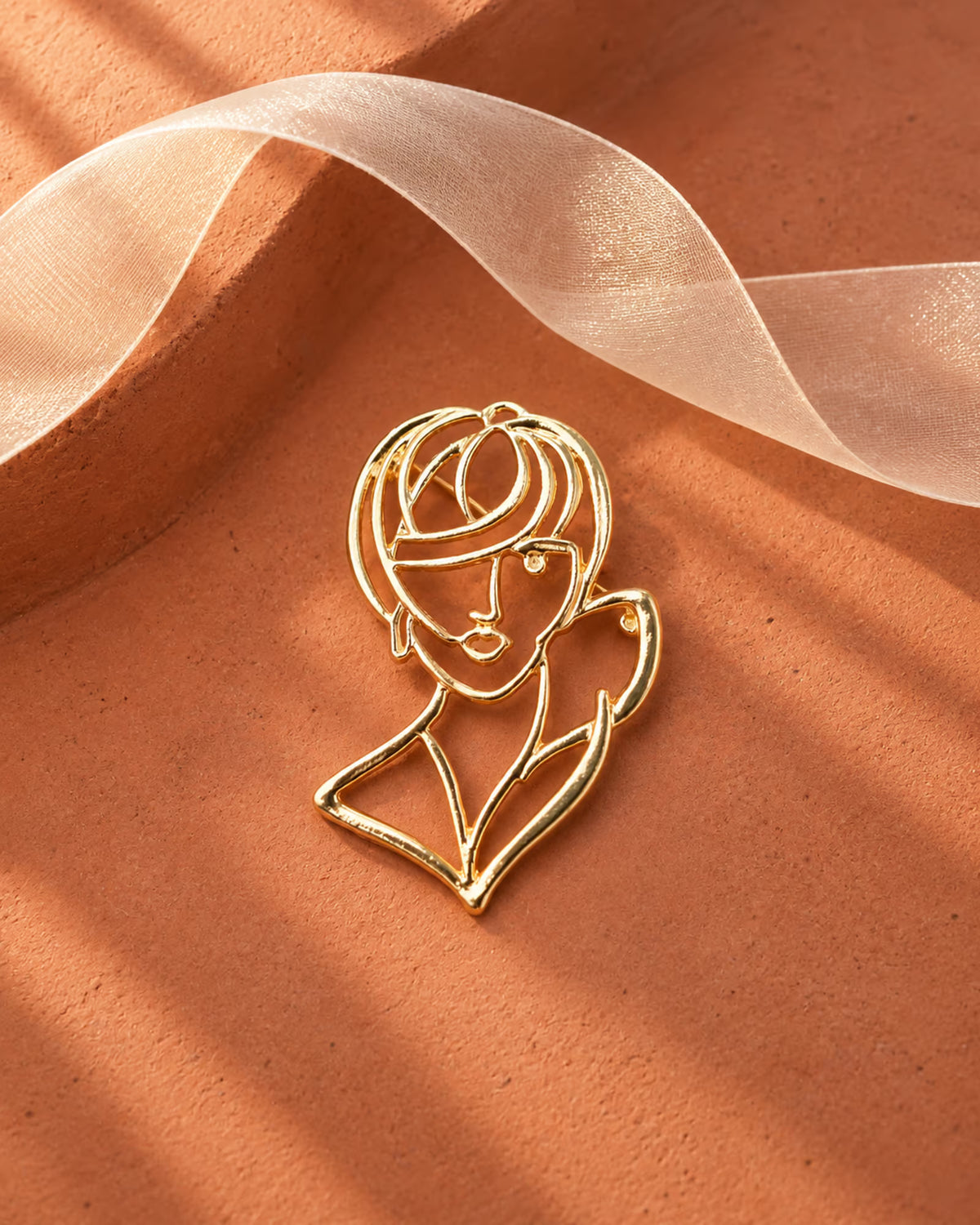 Close-up of Tie Shop Gold Abstract Line Art Face Brooch displayed on a terracotta surface with white organza ribbon. صورة مقربة لـ بروش وجه المرأة الفني الذهبي من تاي شوب على سطح ترابي   وشريط أورجانزا. بروش للحجاب والملابس من تاى شوب 