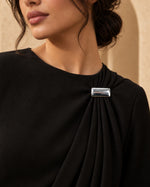 Model wearing a black pleated formal dress styled with the Minimalist Silver Rectangular Brooch from Tie Shop on the shoulder. موديل ترتدي فستان سهرة أسود بطيات أنيقة مزين بـ بروش فضي مستطيل عصري من تاي شوب على الكتف.