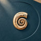 Close-up of Tie Shop Gold Swirl Nautilus Textured Brooch on a dark blue background, an elegant metallic accessory for abayas and dresses. صورة مقربة لـ بروش ذهبي بتصميم الحلزون المموج من تاي شوب على خلفية كحلية، اكسسوار معدني أنيق للعبايات والفساتين.
