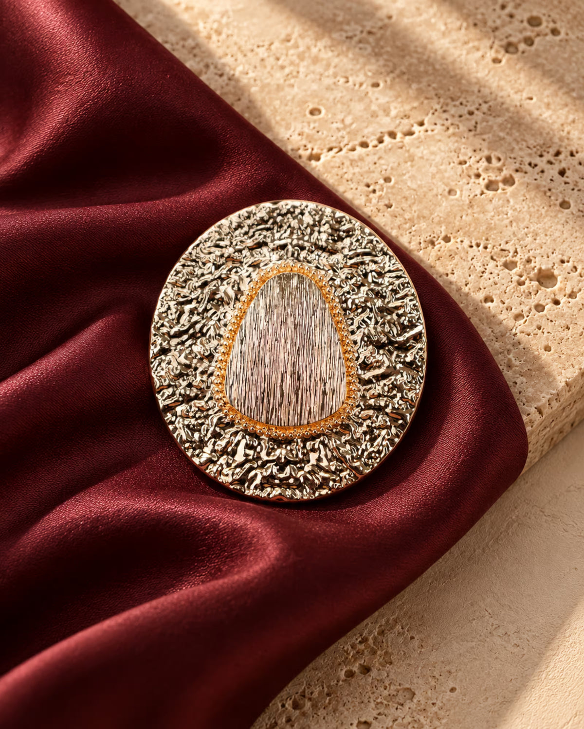 Close-up of Tie Shop Vintage Oval Textured Brooch with golden hammered frame and center stone on burgundy satin, an elegant accessory for abayas and dresses. صورة مقربة لـ بروش بيضاوي بتصميم ريترو محفور بلون ذهبي من تاي شوب على قماش ستان نبيتي، اكسسوار راقي للعبايات والفساتين.