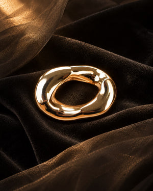 Close-up of Tie Shop Chunky Gold Organic Ring Brooch placed on dark brown textured fabric, a luxury statement metallic pin. صورة مقربة لـ بروش سميك ذهبي بتصميم حلقة عضوية مموجة من تاي شوب على خلفية قماشية دافئة، دبوس معدني فاخر بروش تاى شوب لإطلالة أنيقة مع الحجاب