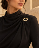 Model wearing a black high-neck draped gown styled with Tie Shop Chunky Gold Organic Ring Brooch on the shoulder, a modern chic fashion brooch. موديل ترتدي فستان أسود أنيق بياقة مرتفعة مزين بـ بروش سميك ذهبي بتصميم حلقة مموجة من تاي شوب، لمسة راقية للملابس.اكتشفي أناقة بروش تاى شوب مع الحجاب والملابس
