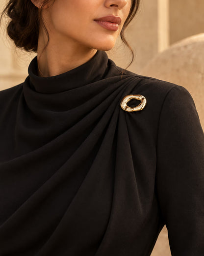 Model wearing a black high-neck draped gown styled with Tie Shop Chunky Gold Organic Ring Brooch on the shoulder, a modern chic fashion brooch. موديل ترتدي فستان أسود أنيق بياقة مرتفعة مزين بـ بروش سميك ذهبي بتصميم حلقة مموجة من تاي شوب، لمسة راقية للملابس.اكتشفي أناقة بروش تاى شوب مع الحجاب والملابس