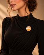 Model wearing an elegant black draped dress styled with Tie Shop Smooth Gold Disc Brooch on the chest, a luxury minimalist brooch accessory for women's hijab and outfits. موديل ترتدي فستان أسود أنيق مزين بـ بروش نسائي من تاي شوب بتصميم دائري ذهبي مصقول، إكسسوار أنيق للحجاب والملابس.