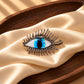 Close-up of Tie Shop Silver Blue Eye Crystal Brooch on beige silk cloth inside a wooden tray, an artistic metallic accessory. صورة مقربة لـ بروش العين الزرقاء الفضي من تاي شوب على قماش حرير بيج وطبق خشبي، اكسسوار معدني فني.